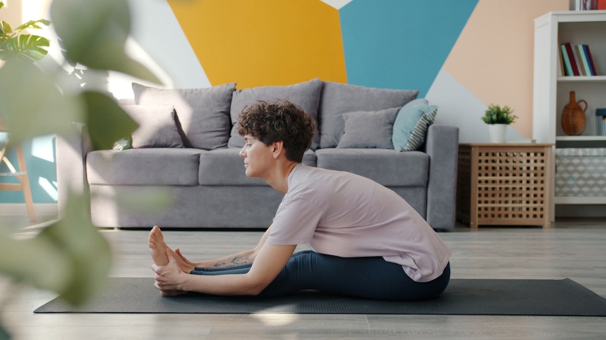 Slow motion of attractive curly-haired woman doing yoga at home bending to legs sitting on mat in cozy apartment enjoying healthy activity. People and lifestyle concept.